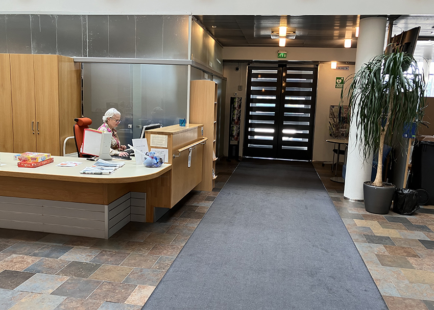 Invalidiliiton esteettömän toimitalon aulassa oleva palvelutiski ja aulan vastaanottovirkailija. Nauvontien puoleinen sisäänkäynti on taustalla.