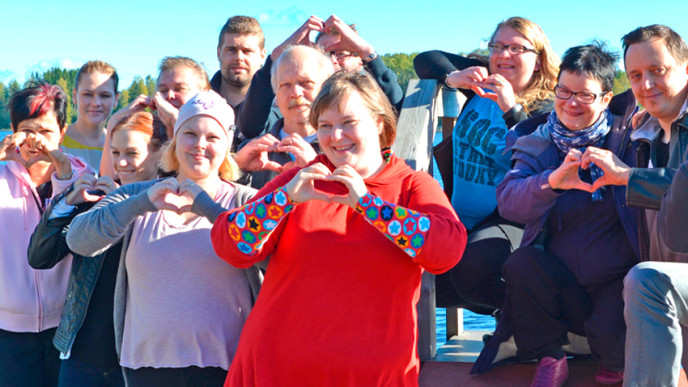 Joukko iloisia ihmisiä seisoo laiturilla järven rannalla kesällä ja tekevät sormillaan sydämen.