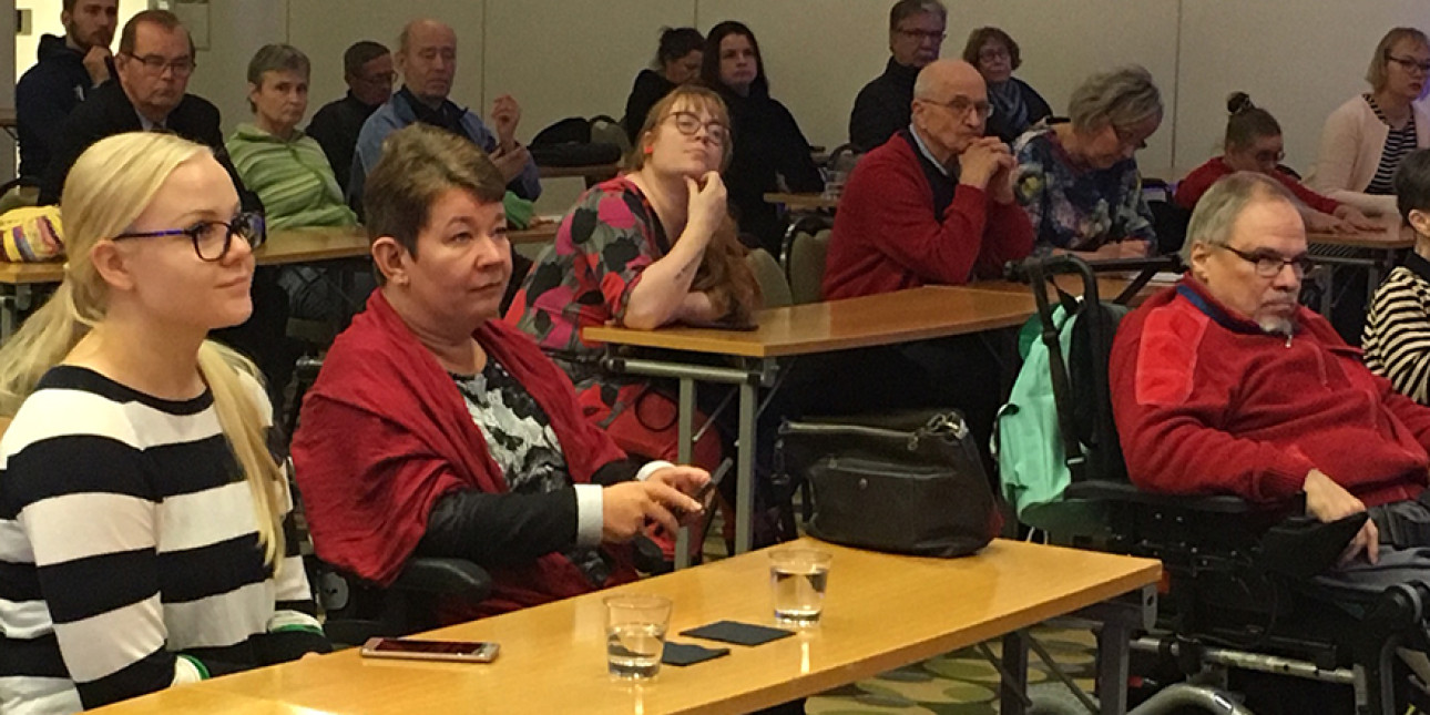 Ihmisiä istuu yleisössä ja kuuntelee puhujaa.