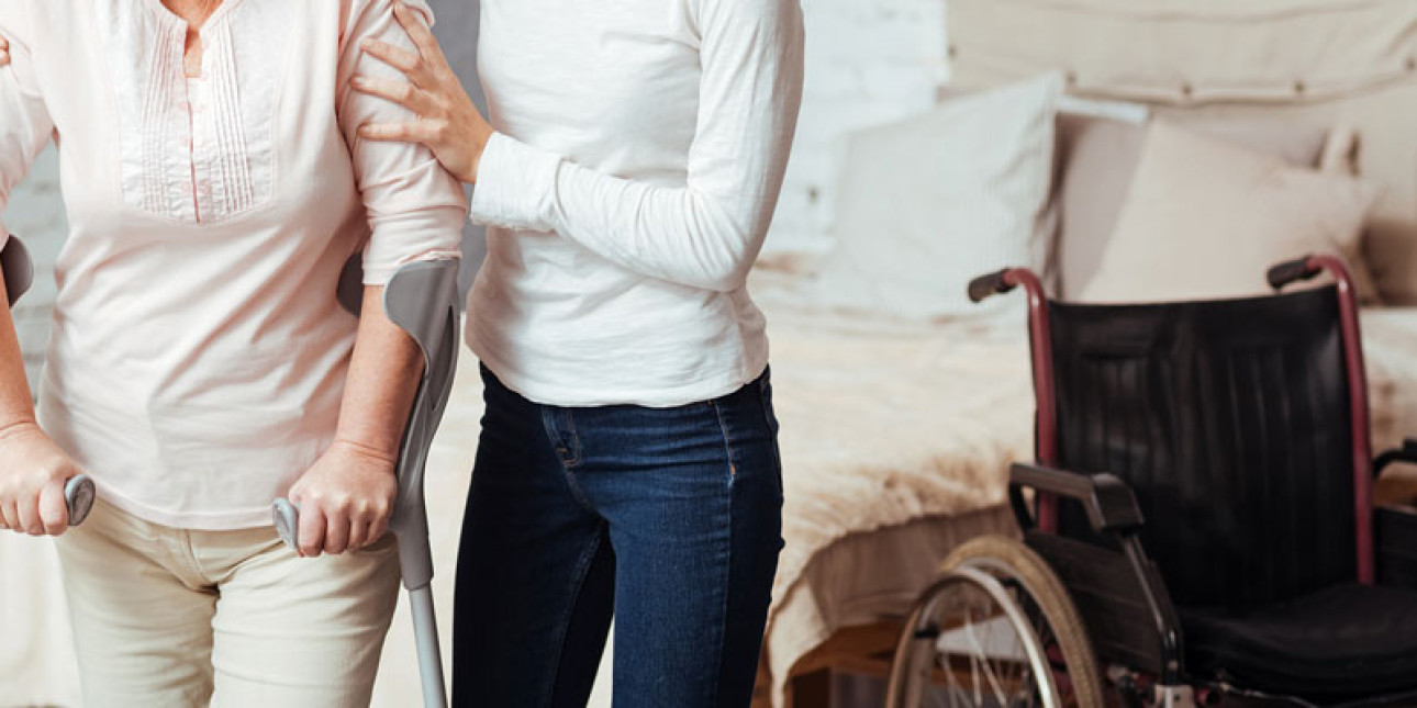 Nuori nainen auttaa vanhempaa naista jolla kyynärsauvat. Taustalla pyörätuoli