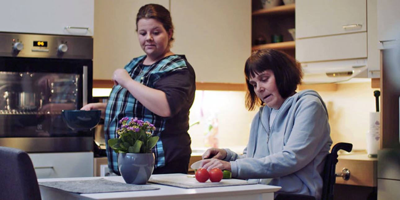 Keittiössä nainen pyörätuolissa leikkaa kurkkua, leikkuulaudalla myös 2 tomaattia. Hänen vieressään seisoo toinen nainen astia kädessä.