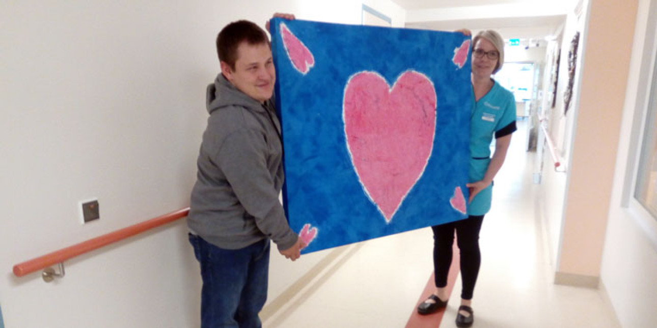 Nuori mies harmaassa hupparissa pitelee kiinni naisen kanssa jolla sininen työasu kangastaulua jossa keskellä iso maalattu vaaleanpunainen sydän ja reunoissa pienemmät sydämet.    