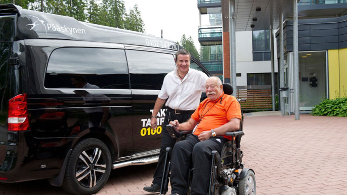 Mieskuljettaja auttaa pyörätuolissa istuvaa miesasiakasta taksiin.