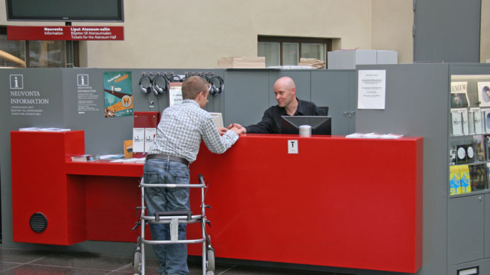 Selin kuvassa oleva rollaattorin kanssa kulkeva nuorimies punaisen neuvontatiskin edessä, jonka takana mies palvelee häntä.