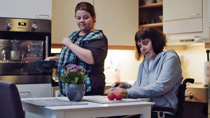 Keittiössä nainen pyörätuolissa leikkaa kurkkua, leikkuulaudalla myös 2 tomaattia. Hänen vieressään seisoo toinen nainen astia kädessä.