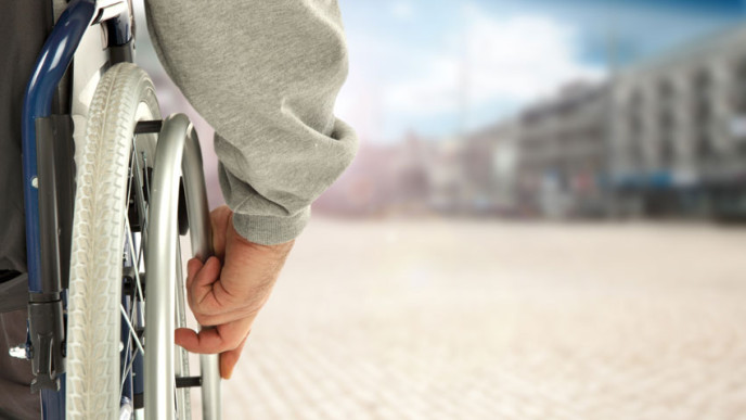 Osa pyörätuolin rengasta ja käsi takaapäin edessä kerrostaloja. Kuvituskuva.