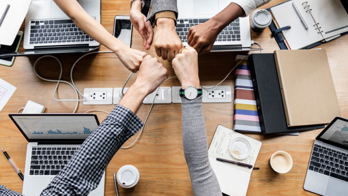 Ihmisiä tietokoneiden ympärillä ja kädet kurottavat yhteen pöydän yli.