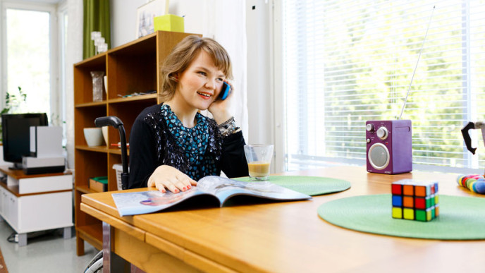 Nuori nainen pyörätuolissa pöydän ääressä puhuu kännykkään. Kuvituskuva.