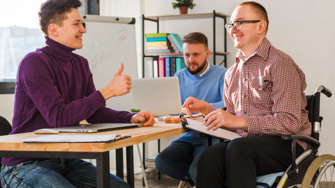 Miehiä istumassa pöydän äärellä, yksi heistä pyörätuolissa. Kuvituskuva.
